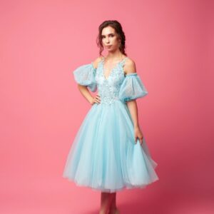A young woman in an airy dress with sleeves and beige shoes poses against an isolated background. A beautiful brunette in a blue ball gown and evening hairstyle holds an airy skirt in her hand