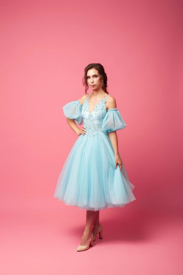 A young woman in an airy dress with sleeves and beige shoes poses against an isolated background. A beautiful brunette in a blue ball gown and evening hairstyle holds an airy skirt in her hand